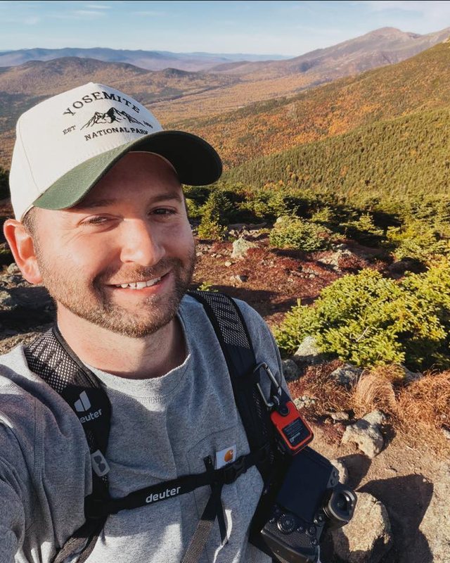 Andrew on a hiking trail with his camera, fall foliage on the hills behind him